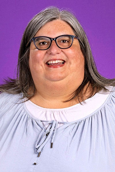 A woman with salt and pepper hair, and wearing glasses and a lavender top, smiles for a formal portrait.