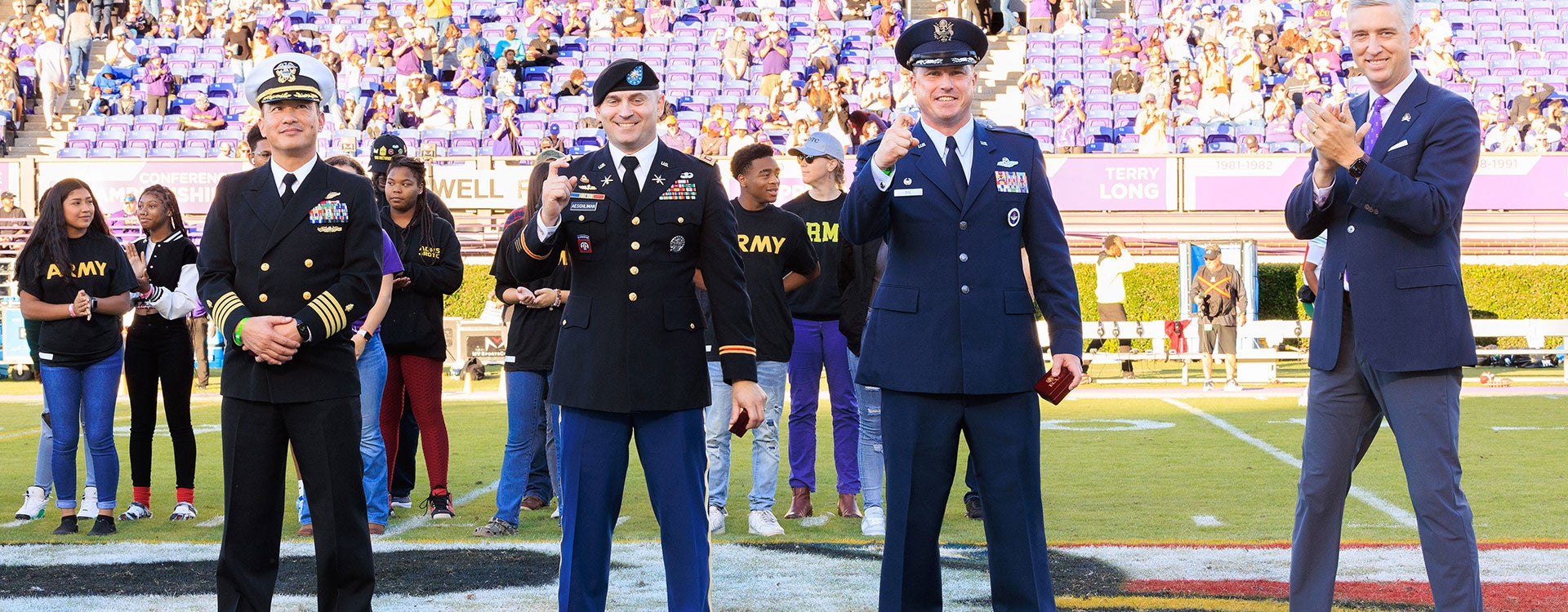 Three men in United States military dress uniforms stand with East Carolina University Chancellor Philip Rogers, who is smiling and clapping, while being recognized on the field at Dowdy-Ficklen Stadium during a football game in 2023. 