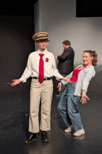 An actor dressed in a shirt, tie, and soldier-style hat stands holding their hands out from their sides. On their right, a woman leans back with her hands out from her sides. They are acting in a production of “Much Ado About Nothing.”