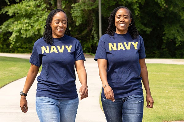 ): Two sisters in blue Navy T-shirts with similar builds and features are photographed smiling and walking on a paved pedestrian path on campus.