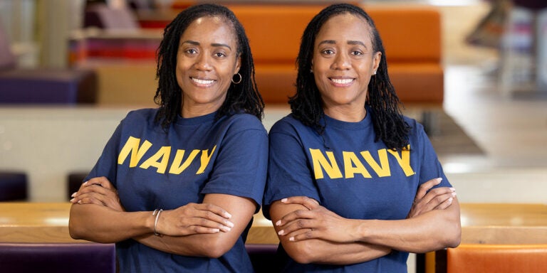 Two sisters in blue Navy T-shirts with similar builds and features stand shoulder to shoulder with their arms crossed, smiling at the camera.