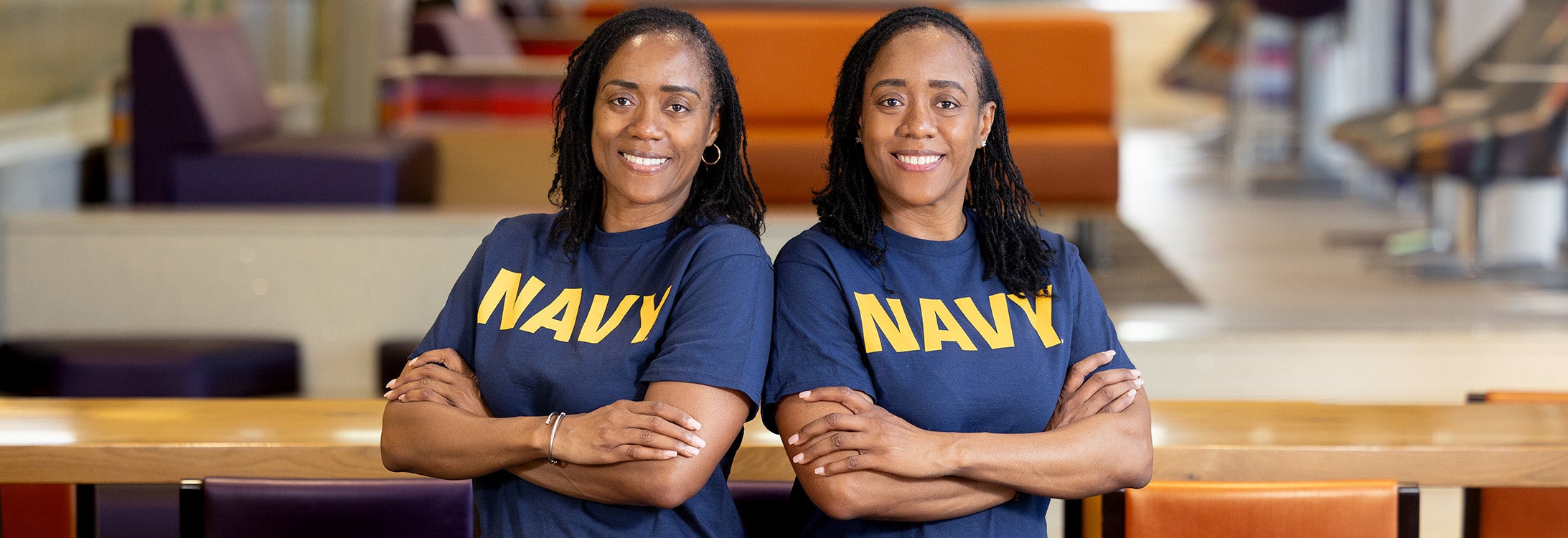 Two sisters in blue Navy T-shirts with similar builds and features stand shoulder to shoulder with their arms crossed, smiling at the camera.