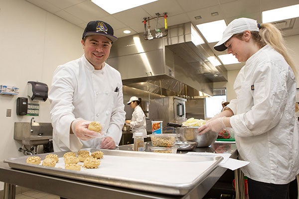 A man and a woman make cookies in a commercial kitchen.