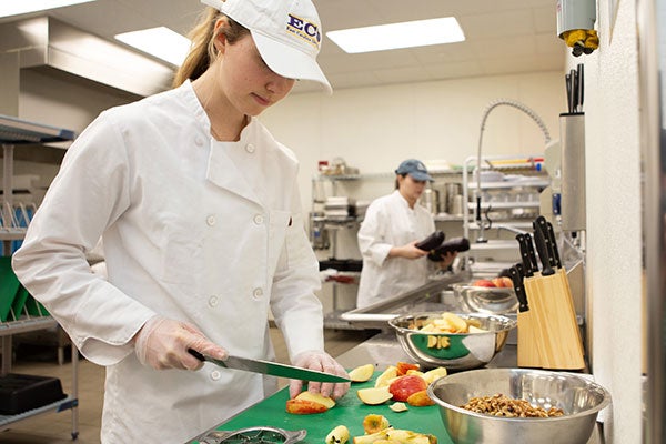 A woman in a white chef’s coat cuts an apple in a commercial kitchen.