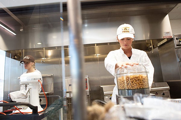 Two women in white coats and baseball hats use kitchen tools to prepare food.