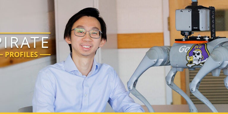 A man wearing glasses and a light blue, collared shirt sits at a desk next to a gray robotic dog that has an East Carolina University Pirate sticker on it.