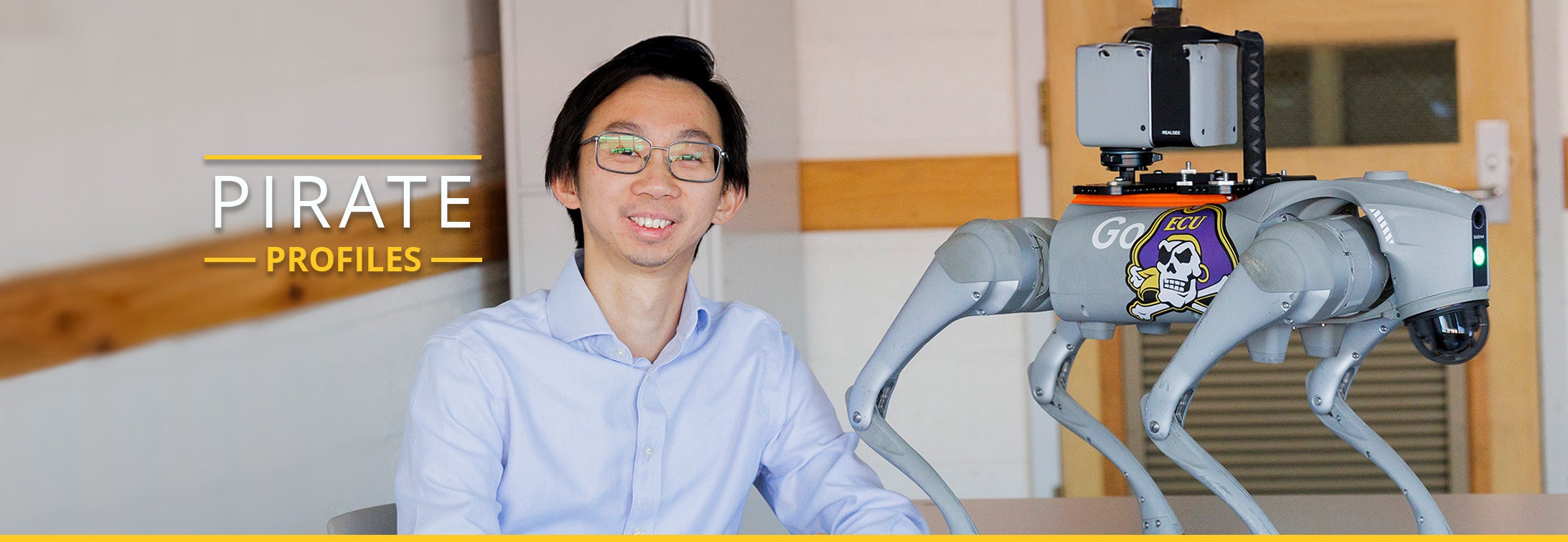 A man wearing glasses and a light blue, collared shirt sits at a desk next to a gray robotic dog that has an East Carolina University Pirate sticker on it.