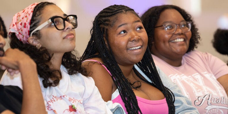 Several students wearing pink and sitting in a row look up at someone on a stage and smile.