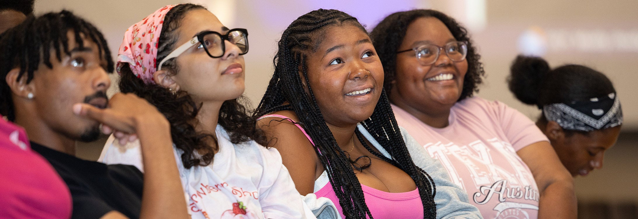 Several students wearing pink and sitting in a row look up at someone on a stage and smile.