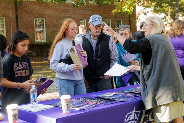 ): A woman stands behind a table pointing while a young woman and older man listen.