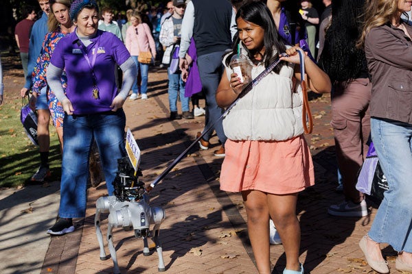 A girl in a white jacket and peach skirt walks a robotic dog.