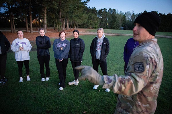 : A man in uniform speaks to a group of women in a field in the early morning light.