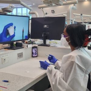 A student dressed in full dental personal protective equipment sits at a workstation in a simulation lab in front of a computer display and holding dental instruments.