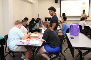 male student stands at table where three students are sitting and placing counting blocks