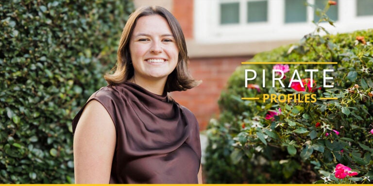 A college age woman wearing a brown top smiles as she sits on East Carolina University’s campus.