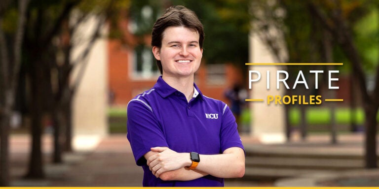 Sam Huffman poses in a purple polo shirt with arms crossed and a smile with blurred trees and Joyner buildings visible in the background.