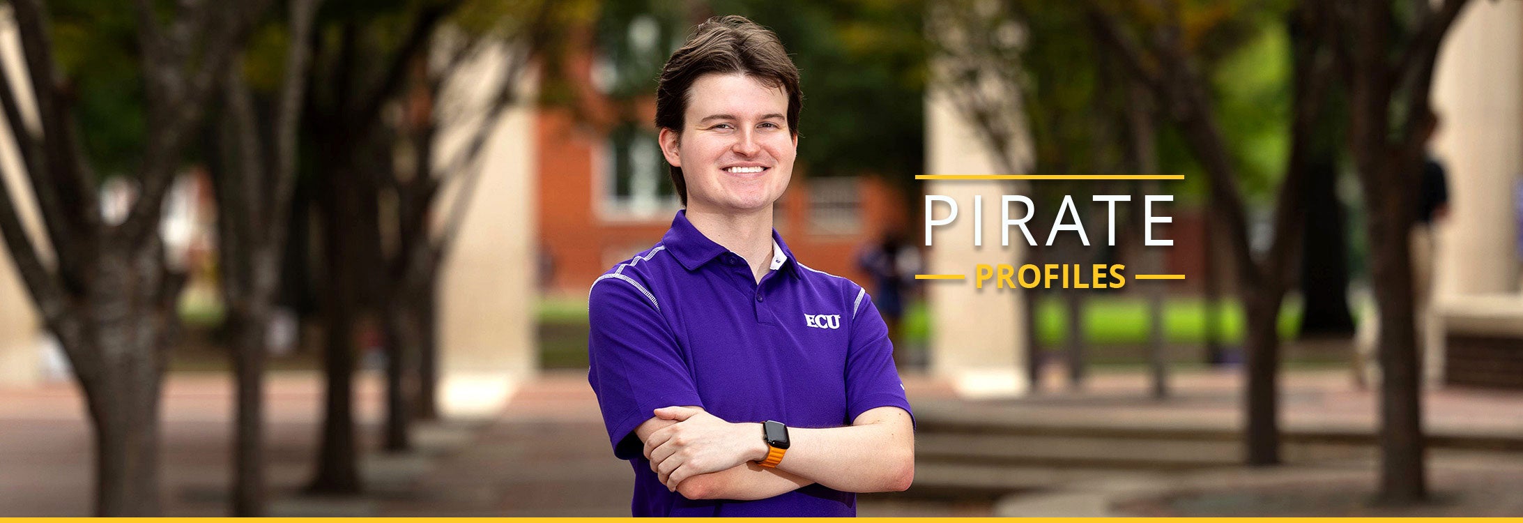 Sam Huffman poses in a purple polo shirt with arms crossed and a smile with blurred trees and Joyner buildings visible in the background.