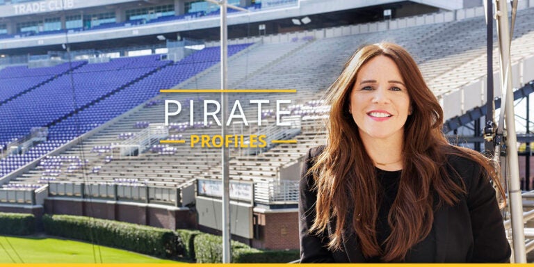 Assistant professor doctor Andrea Buenaño leans against a railing with football stadium bleachers in background.