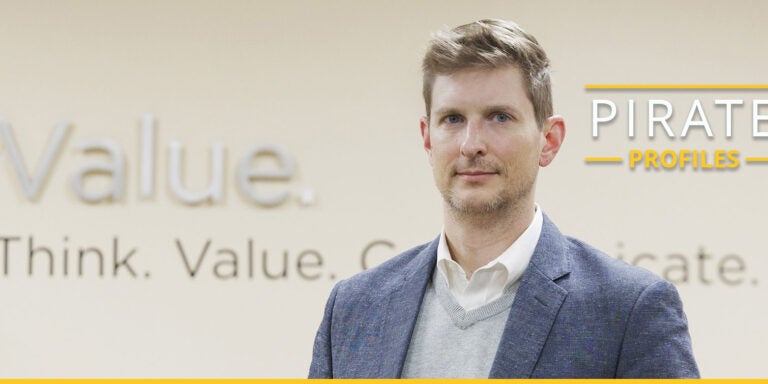 A man in a blue sport jacket and gray sweater stands in front of a wall with words that include value, think and lead.