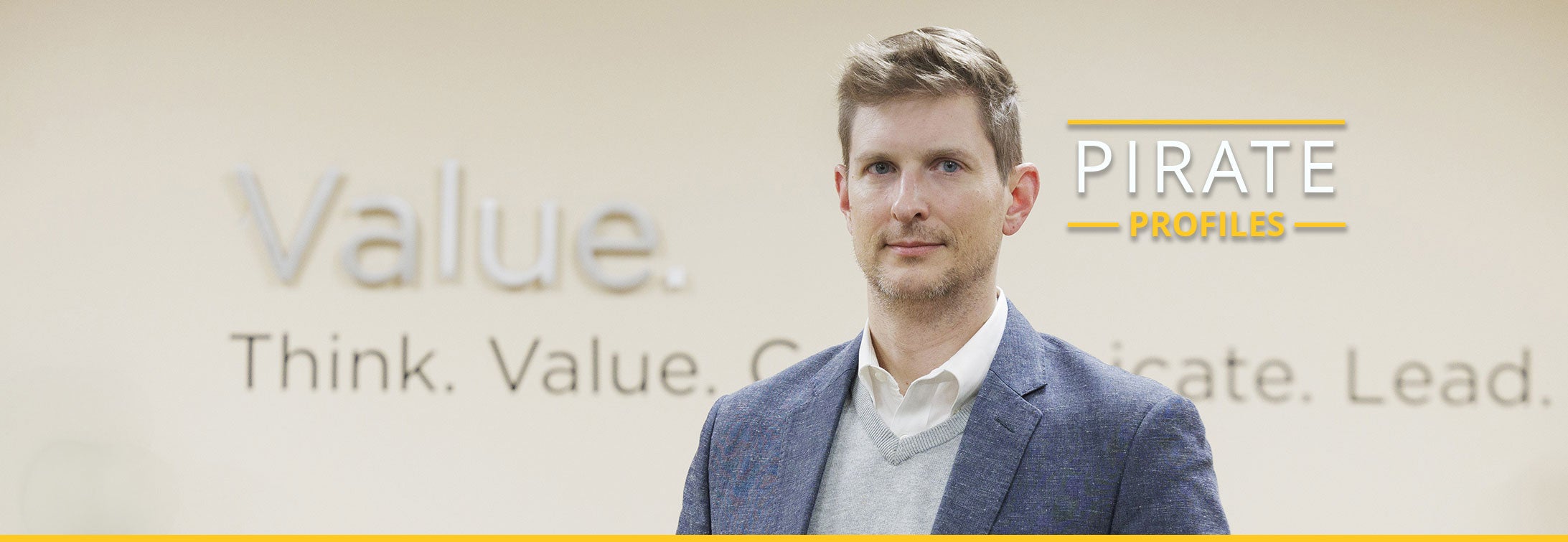 A man in a blue sport jacket and gray sweater stands in front of a wall with words that include value, think and lead.