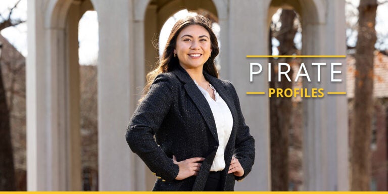 A woman with long dark hair wearing a white blouse under a dark gray tweed jacket stands with her hands on her hips and smiles in front of the Cupola at East Carolina University.