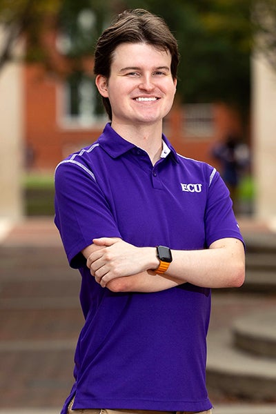 Sam Huffman, wearing a purple ECU polo shirt, adjusts a large circular ring light on a stand, with a green screen in the background.