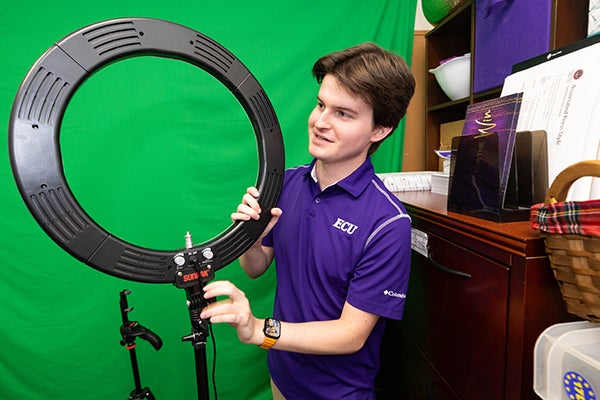 Sam Huffman, wearing a purple ECU polo shirt, adjusts a large circular ring light on a stand, with a green screen in the background.