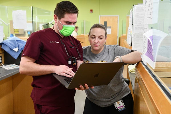 A dental student and faculty member consult, standing with a laptop computer between them as other providers care for patients in the background.