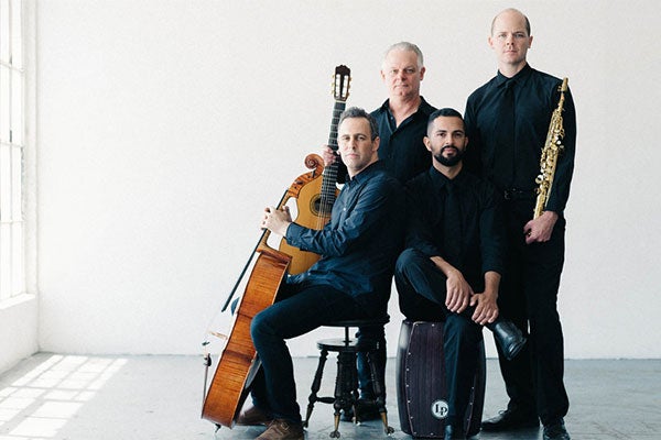 Four male musicians who are part of the music group Quarteto Nuevo pose together in a bright, minimalist setting. Two members are seated in front, with two standing behind them. They hold their instruments: a cello, two guitars or similar string instruments, and what appears to be a woodwind instrument. All are dressed in dark clothing.