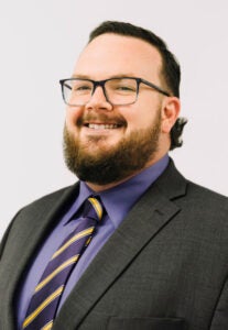 Professional headshot of a man with glasses, short dark hair, and a beard, wearing a dark suit, purple shirt, and striped tie, smiling at the camera against a light background.