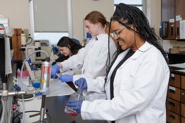 Three smiling female students in lab coats stand at a lab bench with beakers, jars with different colored liquids and a microscope.