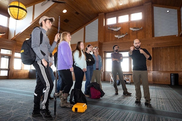 Male professor addresses a group of students by motioning with arms.