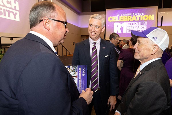 A tall man wearing a suit and purple-striped tie smiles while talking with two other men wearing suits with a large LCD screen in the background showing “A Campuswide Celebration R1 Top-Tier Research University.”