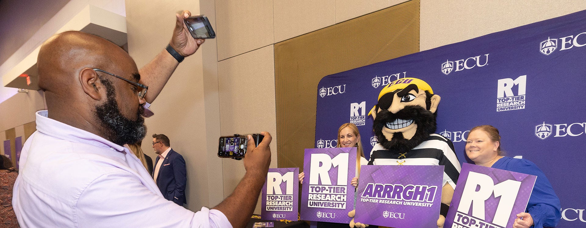A man wearing a purple shirt and khakis takes a photo of two women and PeeDee, East Carolina University’s mascot, holding signs to celebrate the university’s new Research 1 designation.