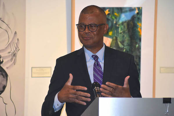 An African American man with glasses in a dark suit stands behind a podium while speaking. 