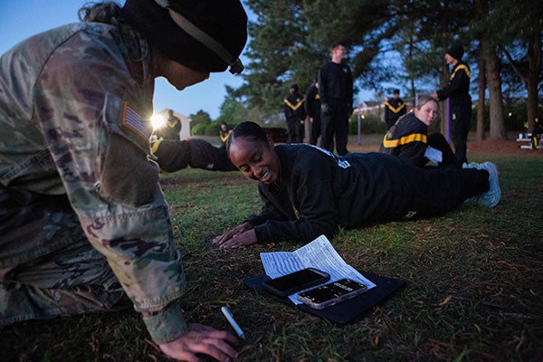 A woman in a black sweatshirt does exercise as another in Army camouflage uniform looks on.