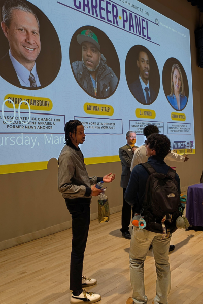 Two college students and two older men stand together, speaking, in front of a projection screen showing four portrait photos and labeled with the text, “Career Panel.”