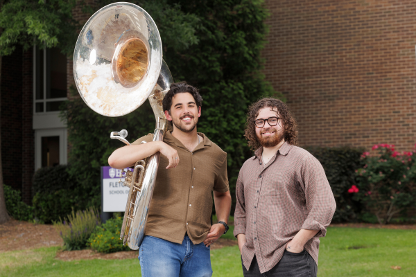Two college students in button-up shirts and jeans stand outside a brick building with landscaped bushes and trees. The student on the left holds a euphonium over his shoulder.