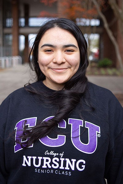 A woman with long dark hair, wearing a black sweatshirt with the East Carolina University logo, stands outside of the College of Nursing building.