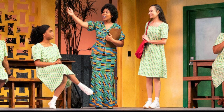 Four female students wearing matching school uniform green and orange polkadot dresses listen to a teacher at a high school during a dress rehearsal for East Carolina University’s production of “School Girls; Or, the African Mean Girls Play.”