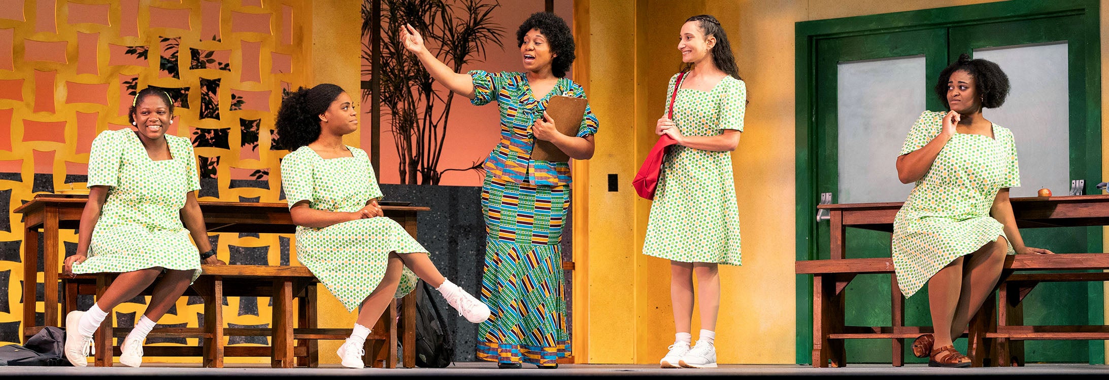 Four female students wearing matching school uniform green and orange polkadot dresses listen to a teacher at a high school during a dress rehearsal for East Carolina University’s production of “School Girls; Or, the African Mean Girls Play.”