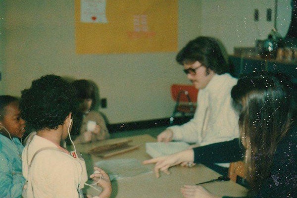 An older photograph shows a man with dark glasses working with children in a classroom setting.