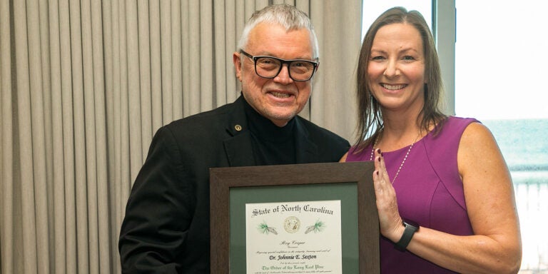 A man in a dark suit and dark rimmed glasses holds a framed certificate alongside a woman in a purple dress. In the background the ocean is visible.