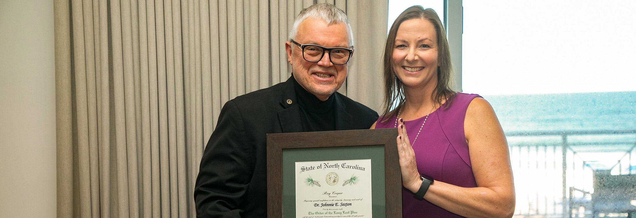 A man in a dark suit and dark rimmed glasses holds a framed certificate alongside a woman in a purple dress. In the background the ocean is visible.