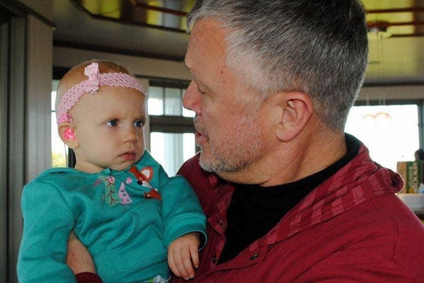 A man in a red sweatshirt holds a young girl in a green outfit and a pink headband.