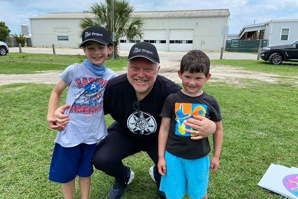 A man in a dark T-shirt and dark baseball hat poses with two young boys.