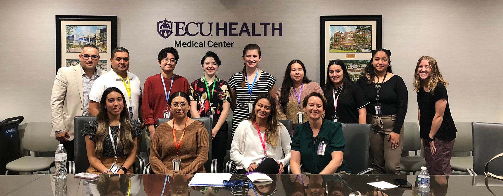 A group of 13 East Carolina University students, faculty and staff sit and stand at a long conference table with two photos and the words ECU Health Medical Center on the wall behind them.