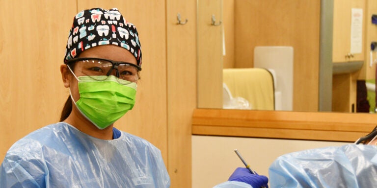 A student pauses in mask and scrub cap as another provider continues working on a patient in a dental suite.