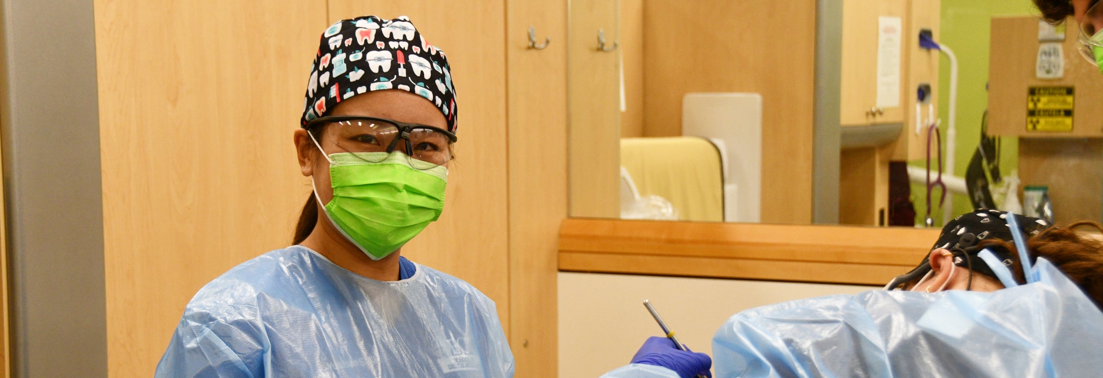 A student pauses in mask and scrub cap as another provider continues working on a patient in a dental suite.