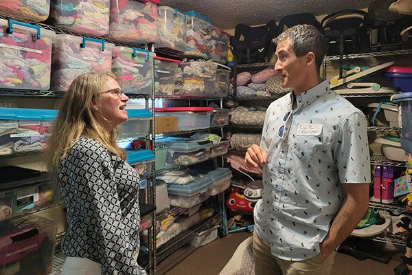 : A man and woman talk with one another while standing in a room filled with donated clothes and children’s goods.